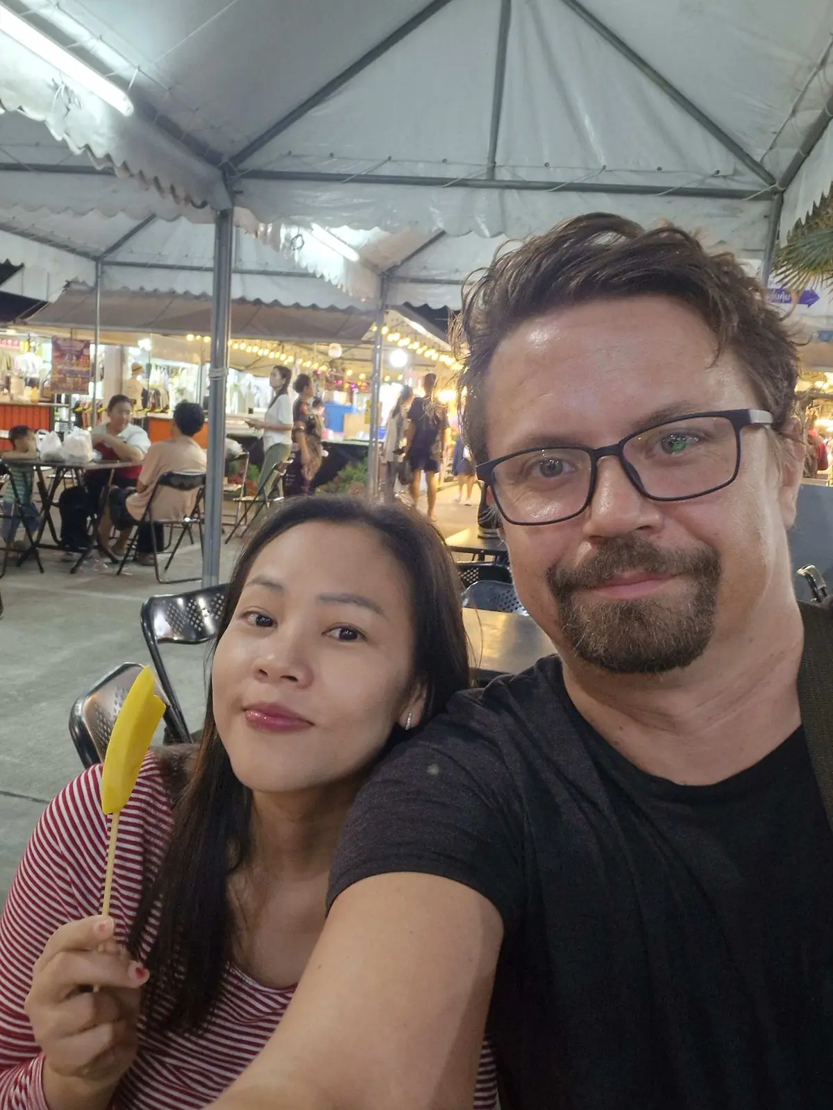 Couple taking a selfie at a night market in Samrong, the woman holding mango on a stick