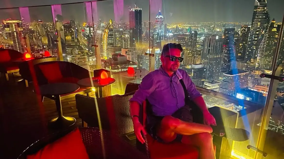 Man at the Red Sky bar at night, red chairs and the lights of Bangkok